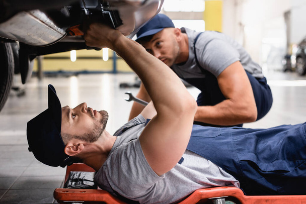 selective focus of mechanic in uniform lying and repairing auto near coworker in car service  - 写真・画像