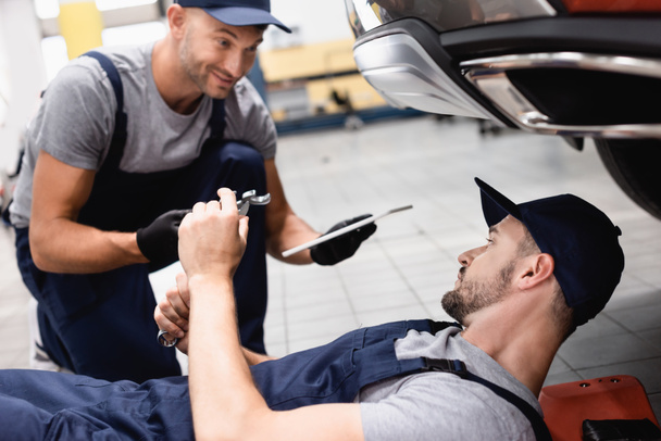 selective focus of mechanic holding digital tablet and giving wrench to coworker in cap repairing car - Фото, зображення