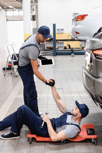 mechanic with digital tablet holding hands with coworker in cap lying near car - Фото, изображение