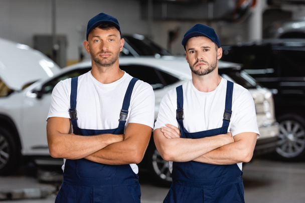 handsome mechanics in uniform and caps standing with crossed arms in car service - Photo, Image