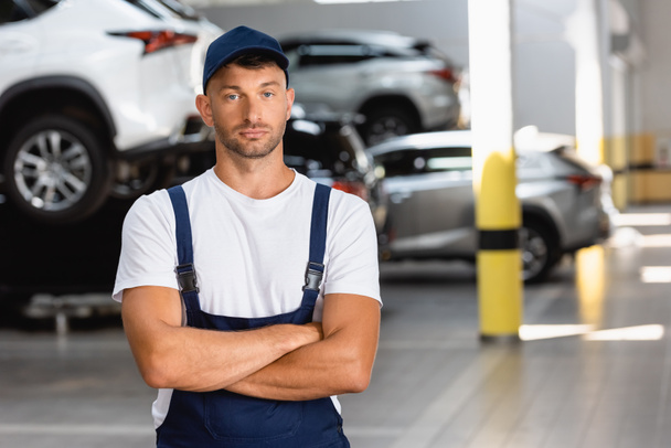 handsome mechanic in uniform and cap standing with crossed arms in service station - Foto, Imagem