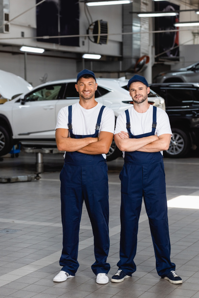 fröhliche Mechaniker in Uniform und Mützen stehen mit verschränkten Armen im Autoservice - Foto, Bild