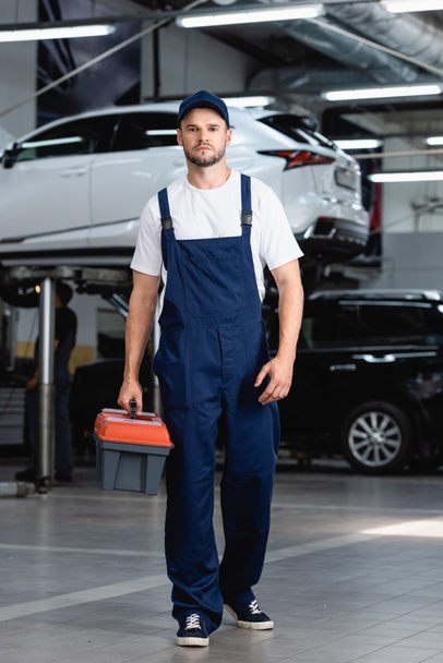 handsome mechanic in overalls and cap holding toolbox in service station - Foto, Bild