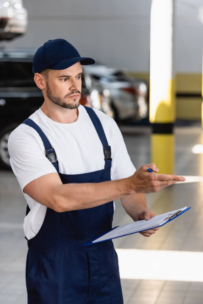 handsome mechanic in uniform and cap pointing with hand while holding clipboard and pen - Fotoğraf, Görsel