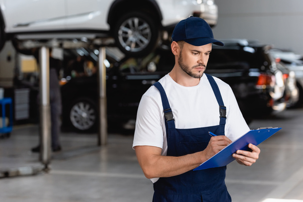 handsome mechanic in overalls and cap writing while holding clipboard - Photo, Image