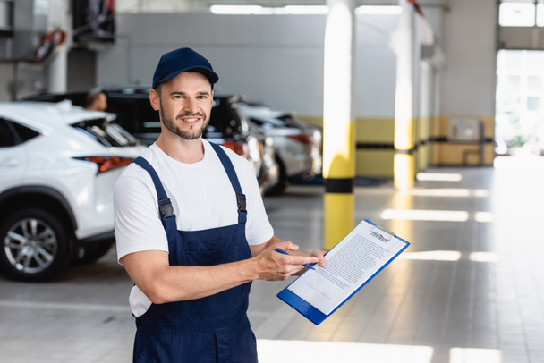 happy mechanic in uniform and cap holding clipboard with contract lettering and pen near cars - Foto, Imagem