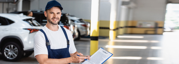 panoramic concept of happy mechanic in uniform and cap holding clipboard with contract lettering and pen near cars - Photo, Image