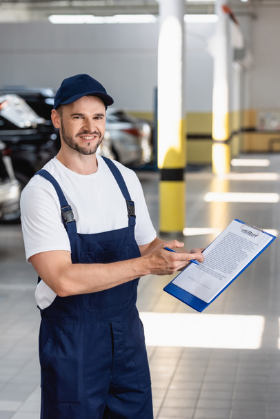 cheerful mechanic in uniform and cap holding clipboard with contract lettering and pen near cars - Photo, Image