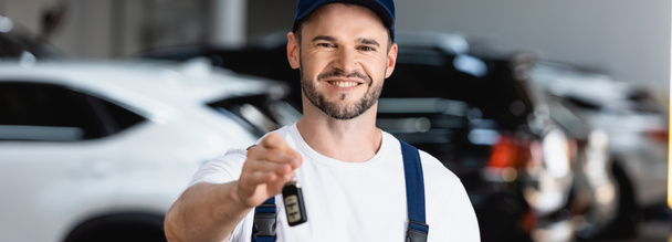horizontal image of cheerful mechanic in uniform and cap holding car key  - Fotoğraf, Görsel