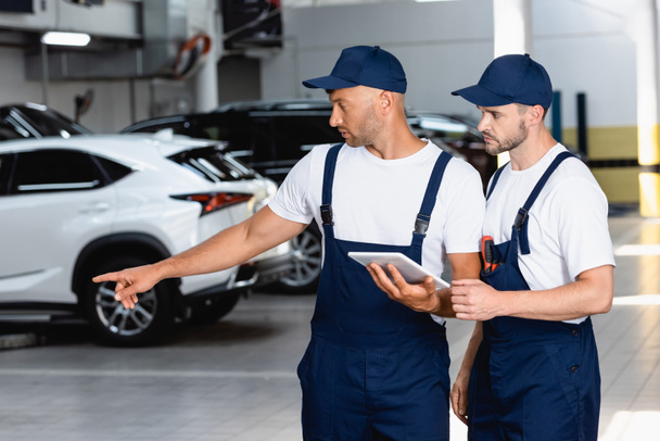 handsome mechanic in uniform holding digital tablet and pointing with finger at car near coworker in workshop  - Photo, Image