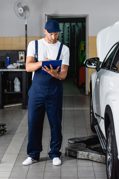 handsome mechanic in cap holding clipboard and writing near car - Фото, зображення