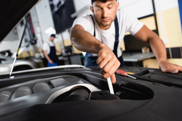 selective focus of handsome mechanic in cap repairing car near coworker in workshop  - Foto, Imagen