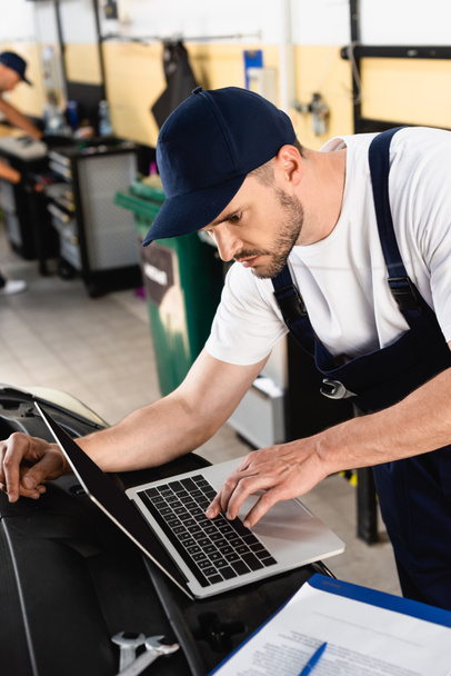 Auto mechanic typing on laptop on car near clipboard and wrenches at service station - Foto, imagen