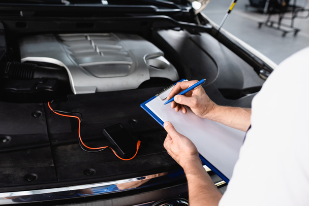 Cropped view of auto mechanic writing on clipboard near open hood of car with multimeter at service station - Fotoğraf, Görsel