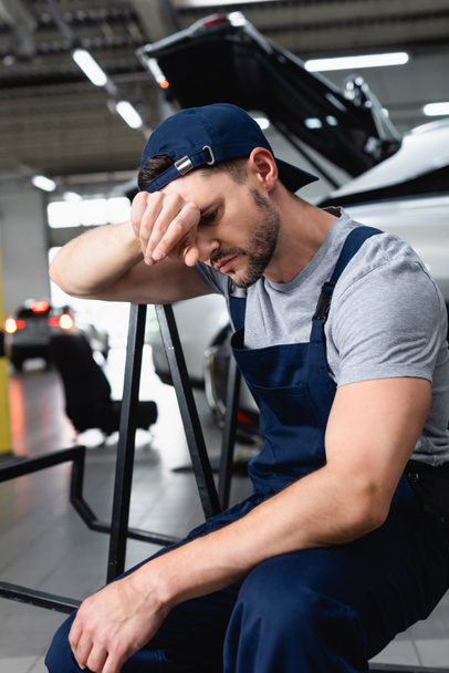 Tired auto mechanic with hand near forehead sitting near cars at service station - 写真・画像
