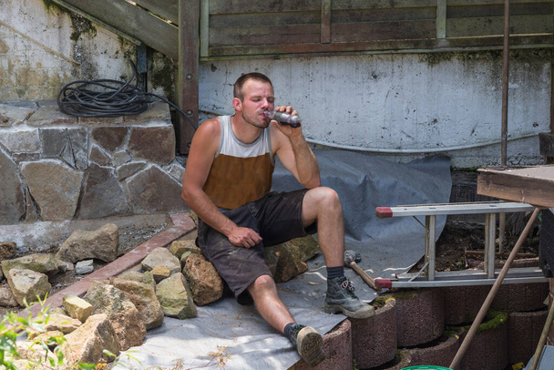 Les artisans en vêtements de travail font une pause. Assis sur le sol d'un chantier de construction tenant une boisson rafraîchissante à la main. - Photo, image