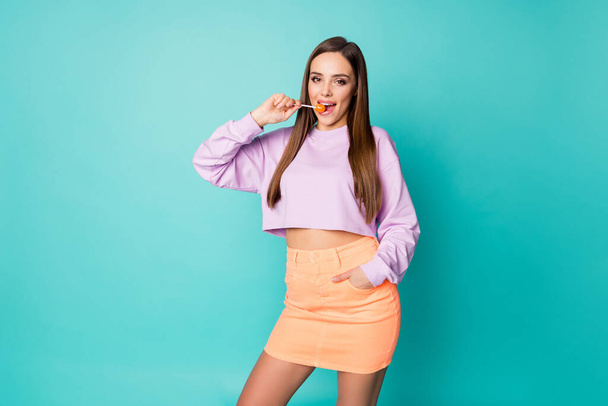 Photo of pretty tender lady hold lollipop hands good mood sweets addicted person lick bite tasty candy wear cropped pullover orange short skirt isolated teal color background - 写真・画像