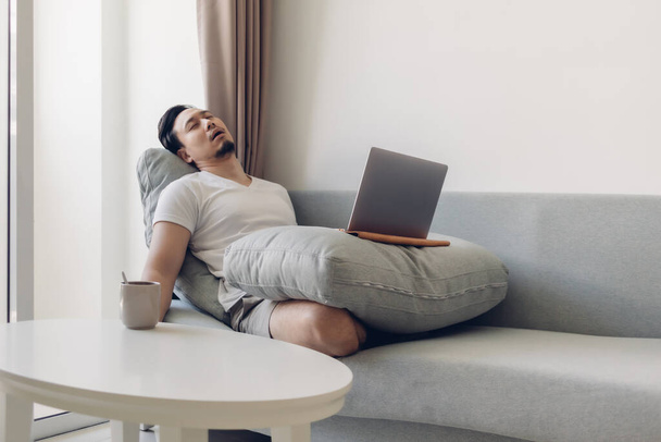 Sleepy Asian man working on his laptop on the sofa. - Фото, изображение