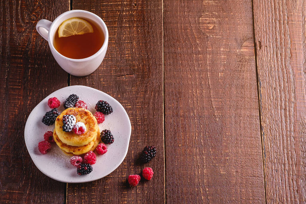 Cottage cheese pancakes, curd fritters dessert with raspberry and blackberry berries in plate near to hot tea cup with lemon slice on dark brown wooden background, angle view copy space - Φωτογραφία, εικόνα