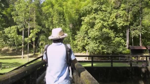 Азиатская туристка в соломенной шляпе и рюкзаке идет по старому бетонному мосту через плотину и фотографируется с помощью мобильного телефона. Путешественник наслаждается провести время в лесном парке под солнечным светом в течение лета. Провинция Пханг Нга. Таиланд. - Кадры, видео