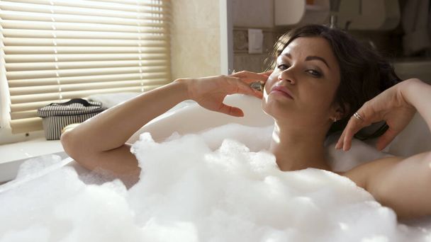 Молодая счастливая женщина в ванной с белой пеной. Видео. Портрет брюнетки с вьющимися волосами и макияжем на лице, принимающей пенную ванну дома. - Фото, изображение