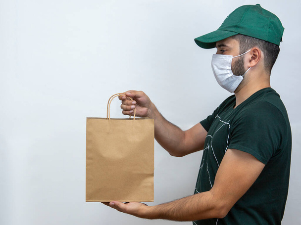 Entrega de mercancías. Entrega en línea del pedido, al trabajo, a la oficina. Repartidor de máscara de paquete en gorra verde y camiseta. Coronavirus. COVID-19
 - Foto, Imagen
