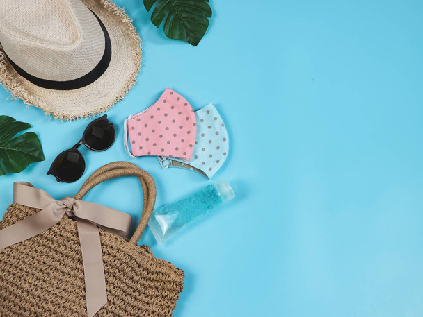 COVID-19 preventie, reizen en nieuw normaal concept, bovenaanzicht van geweven tas met ontsmettingsgel, roze en blauwe polka dot stof gezicht maskers en vrouwen vakantie accessoires op blauwe achtergrond. - Foto, afbeelding