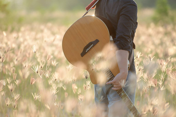 Detail muže hrajícího na kytaru, hudebníka hrajícího na akustickou kytaru, koncept západu slunce na zamlžené louce v letním večeru a květinové pole na slunečním pozadí. - Fotografie, Obrázek
