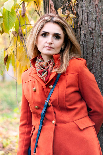 beautiful concentrated woman with long hair in orange coat looking at camera in front of tree with colorful foliage - Foto, Imagem