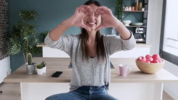 Video of pretty young woman looking at camera while doing heart shape with her hands in the kitchen at home. - Πλάνα, βίντεο