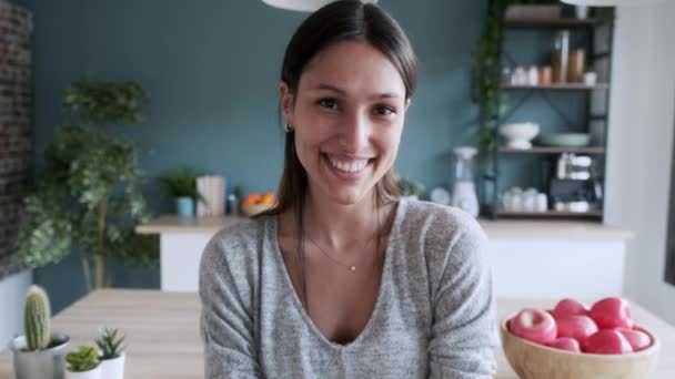 Video of smiling young woman looking at camera while sitting on the stool in the kitchen at home. - Video