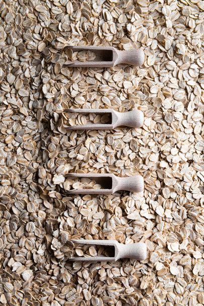 Four wooden scoops with flakes of rolled oats in them. Row of scoops is on layer of rolled oats. Source of vegetable protein. Healthy and sport eating concept background. Vertical orientation. - Photo, Image