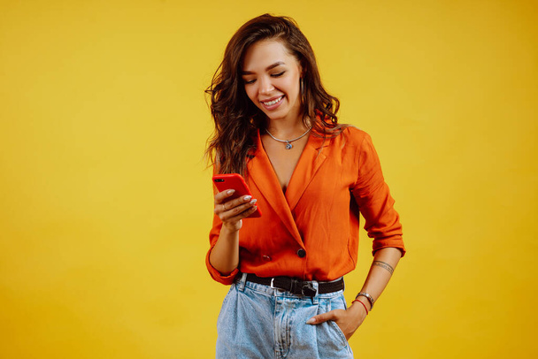 Cheerful young brunette girl with phone posing on yellow background. Fashion and lifestyle concept. Technology concept. - Foto, imagen