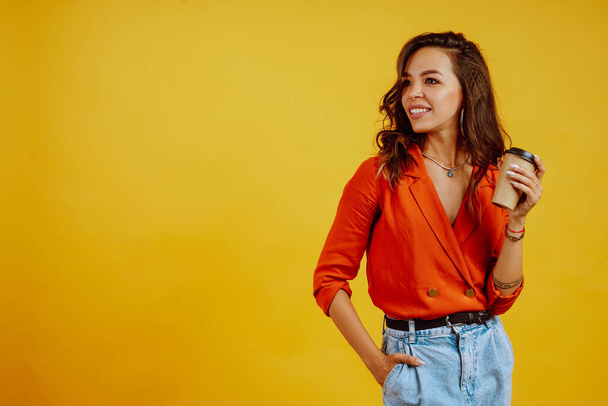 Young woman with coffee posing on yellow background. Portrait of young girl wears a carrot blouse and jeans. Fashion and lifestyle concept. - Φωτογραφία, εικόνα