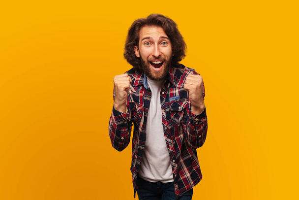 Happy man in plaid shirt celebrating victory clenching his fists for joy on isolated yellow background - 写真・画像