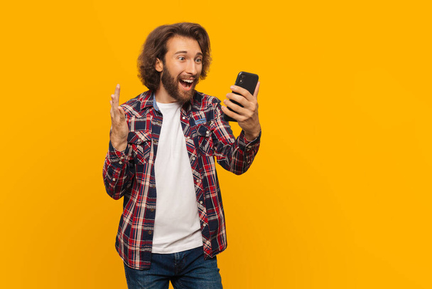 Happy mma man with curly hair in a plaid shirt is very happy about victory looks into a mobile phone and shouts with delight on isolated yellow background - Фото, зображення