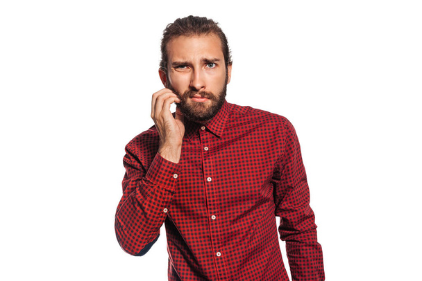 Serious young man in a red plaid shirt with a beard touches his beard with a hand and looks at the camera, isolated on white background - Fotó, kép