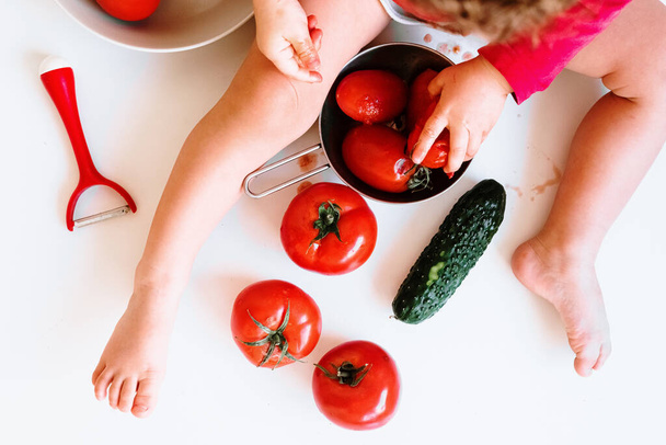 Een blootsvoets baby speelt bij het bijten van rode tomaten en het insmeren van zijn sap. - Foto, afbeelding