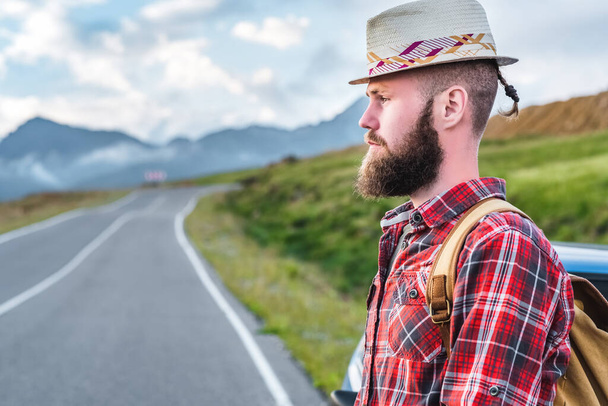 Handsome young man in a straw hat and a plaid shirt, stands on the road, against the backdrop of beautiful mountains. The concept of freedom and hitchhiking - Фото, зображення