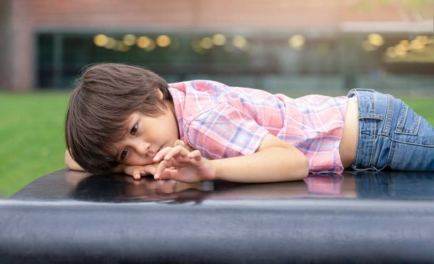 rajattu laukaus yksinäinen pieni poika makaa bence puistossa, Kid tylsistynyt kasvot sormilla kirjallisesti jotain, Surullinen lapsi leikkii omalla ulkona, onneton lapset käsite - Valokuva, kuva