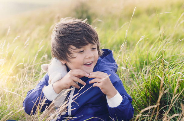 Retrato de niño feliz mostrando juguete de perro mullido en su mano sentado en la hierba en el campo de la granja, Niño con la cara sonriente jugando al aire libre con su juguete en el prado en la colina, concepto de niños positivos
 - Foto, imagen