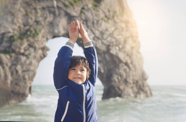 Portarit of Cute kid boy doing yoga standing by the sea with blurry Durdle Door background. Healthy child practicing Yoga vrikshasana tree pose on the beach with morning light, Healthy lifestyle or Yoga for kids concept. - Photo, Image