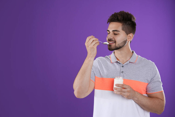 Happy young man eating tasty yogurt on purple background. Space for text - Foto, Bild