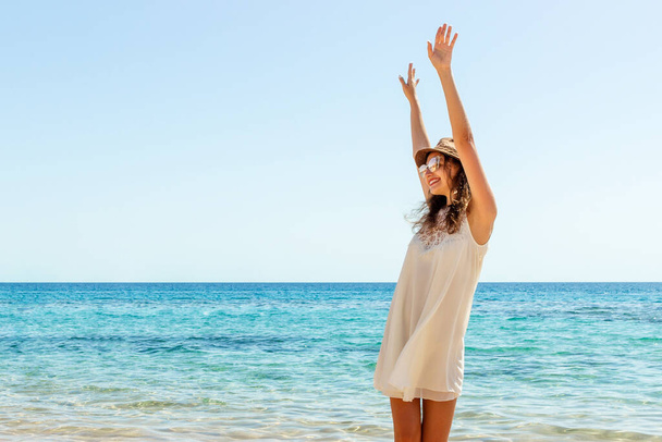 Obraz mladé ženy, oblečené v bílých šatech a hnědém klobouku, stojící na pláži a rozestavěné. Dívka si užívá svobodu a čerstvý vzduch. Pocit volnosti koncept. - Fotografie, Obrázek