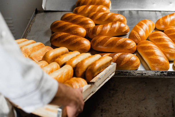 Um padeiro em uma padaria empilha pão quente fresco em uma caixa de madeira. Produção de pão industrial
 - Foto, Imagem