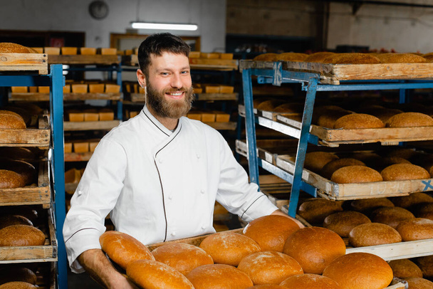 A baker holds a tray with fresh hot bread in his hands against the background of shelves with fresh bread in a bakery. Industrial bread production - Fotó, kép
