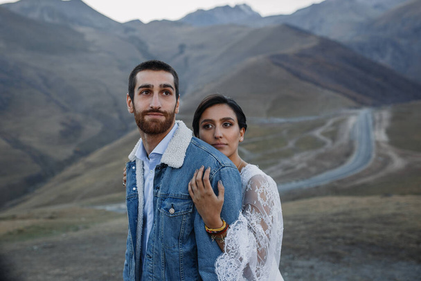 Jeune couple géorgien posant dans les montagnes. Mariage Boho en Géorgie. Soleil couchant du soir. Blouson jeans bleu - Photo, image