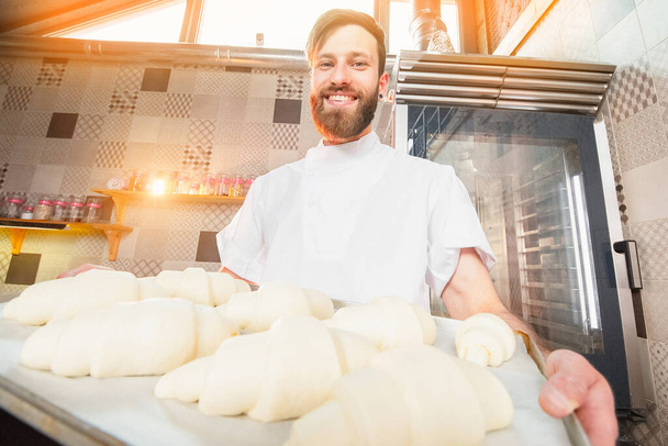 Un panadero joven y guapo sostiene cruasanes crudos de masa blanca en las manos de un horno. Productos semielaborados a partir de masa. - Foto, imagen