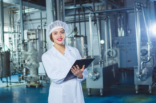 A young beautiful girl in white overalls makes notes in a tablet on the background of equipment of a food processing plant. Quality control in production. - Foto, afbeelding