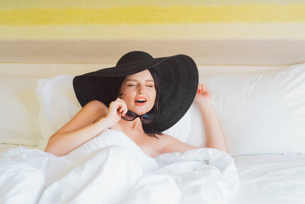 A young girl in a big hat and sunglasses lies in bed. Morning on vacation. Girl woke up in glasses and hat. - Foto, imagen